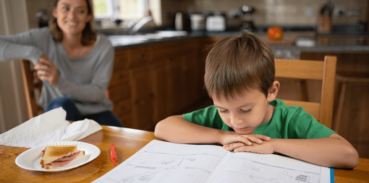 Child studying at a table with an adult in the background.